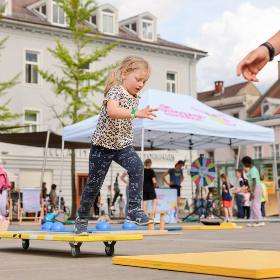 Ein junges Mädchen balanciert auf einem Skateboard an einem sonnigen Tag auf einem Außenbereich mit Menschen um sie herum und einem Zelt.