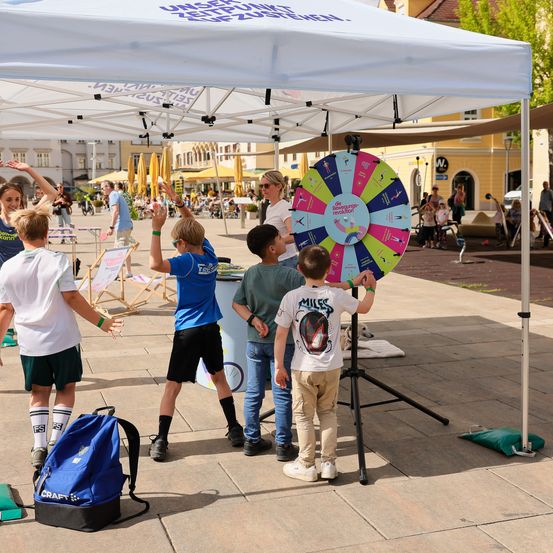 Kinder spielen ein Spiel unter einem weißen Zelt auf einem Stadtplatz. Ein großes Rad mit verschiedenen Farben und Text ist zu sehen. Einige Leute stehen in der Nähe, manche mit Rucksäcken.