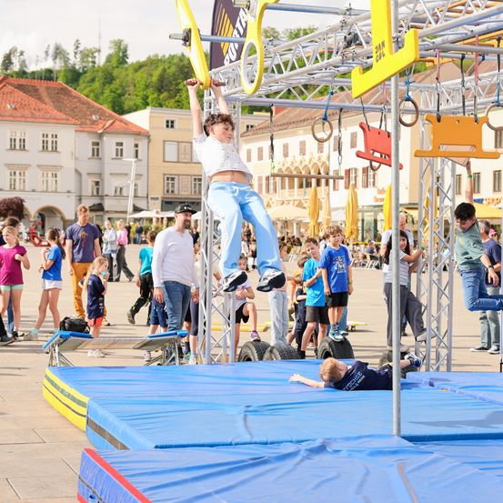 Ein junger Junge hängt an einer Hindernisparcours-Struktur im Freien. Mehrere Leute beobachten und einige gehen herum. Gebäude und Bäume sind im Hintergrund.