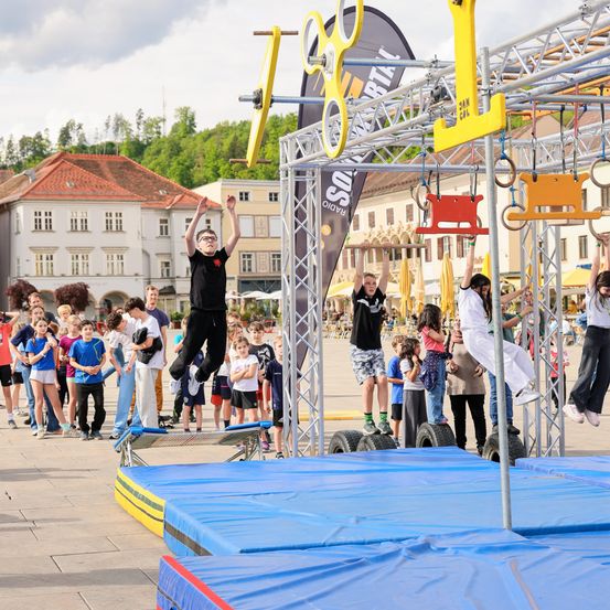 Teilnehmer einer Hindernisparcours-Herausforderung im Freien, umgeben von einer Menschenmenge. Zwei Personen hängen an einem Balken, einer springt von der Plattform ab.