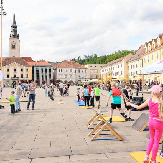 Eine Gruppe von Menschen nimmt an einem Hindernisparcours auf einem Stadtplatz teil. Der Parcours ist mit verschiedenen Hürden aufgebaut. Gebäude mit roten Dächern umgeben den Bereich.