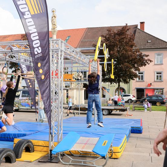 Zwei Frauen auf einem Klettergerüst, eine hält Ringe, die andere hängt an Ringen, auf einer blauen Matte. Dahinter ein Banner und eine Statue. Autos und Gebäude im Hintergrund.