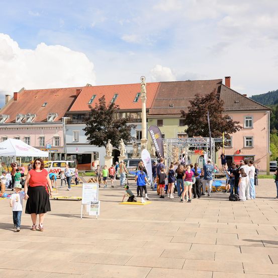 Eine große Gruppe von Menschen auf einem Stadtplatz mit einem Denkmal und mehreren Gebäuden im Hintergrund. Viele Kinder sind anwesend, und eine Frau und ein Kind gehen im Vordergrund.