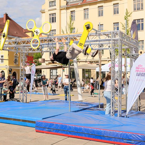 Ein Mann führt einen Stunt auf einem Metallrahmen mit gelben Spinnern auf einem öffentlichen Platz vor, während die Leute von den Seiten zuschauen und Gebäude im Hintergrund stehen.