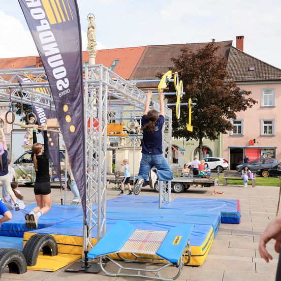 Eine Gruppe von Menschen trainiert auf einer blau-gelben Plattform im Freien. Ein großes Banner mit Text steht in der Nähe.