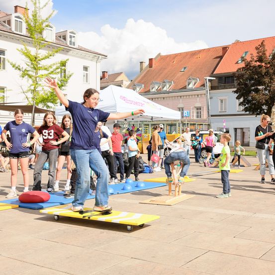 Eine Gruppe von Menschen steht herum und beobachtet zwei Kinder, die auf Brettern in einem Stadtplatz balancieren. Eine Person balanciert auf einem Skateboard, während eine andere einen Rückwärtssalto auf einer Plattform ausführt.