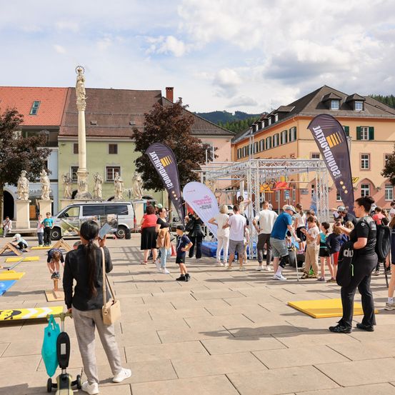 Eine große Gruppe von Menschen versammelt sich auf einem Stadtplatz mit Strukturen, Bannern und Statuen. Einige beobachten, während andere Aktivitäten durchführen. Im Hintergrund sind Fahrzeuge und Bäume sichtbar.