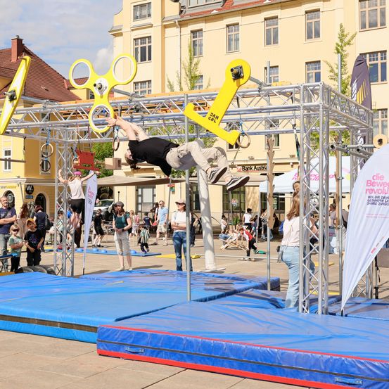Ein Mann führt einen Stunt an einer Metallkonstruktion im Freien aus. Es gibt Zuschauer.