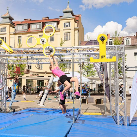 Ein Mädchen in Pink klettert auf einem gelben Gerät, während andere im Stadtplatz zuschauen. Die Umgebung umfasst Gebäude und Bäume.