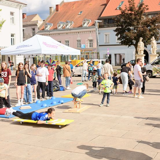 Eine Gruppe von Kindern ist draußen bei einer körperlichen Aktivität, in verschiedenen Sportbekleidungen. In der Nähe beobachten Erwachsene. Im Hintergrund sind Gebäude und ein Zelt zu sehen.