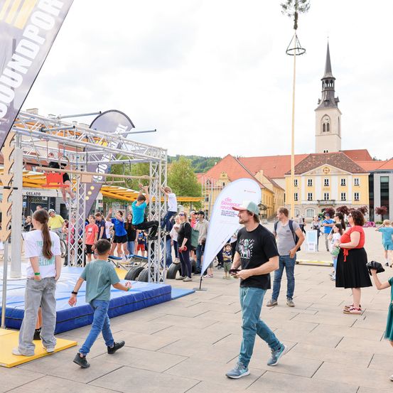 Ein belebter Stadtplatz mit Menschen, die eine Hindernisbahn genießen, einer Kirche und Gebäuden im Hintergrund. Ein Mann geht an einem Mädchen vorbei und ein Junge beobachtet eine Person auf einer Schaukel.