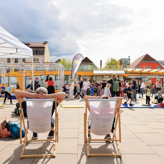 Zwei Personen entspannen in Stühlen auf einer Freiluft-Plaza, mit einer Menge spielender Kinder in der Nähe. Im Hintergrund sind Gebäude, ein Zelt und eine große Struktur zu sehen.