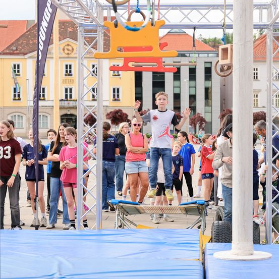 Ein Junge springt auf einer Plattform mit Kletterringen vor einer Menge. Gebäude und Bäume sind im Hintergrund.