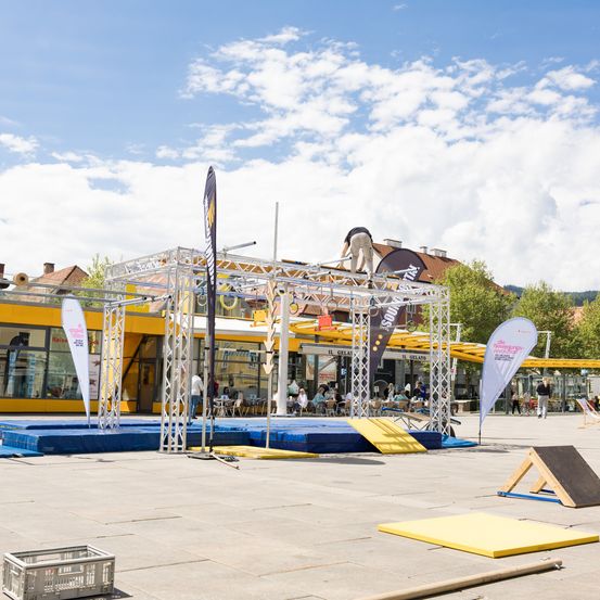 Eine Metallstruktur für Outdoor-Aktivitäten steht auf einem Stadtplatz. Eine Person klettert auf die Struktur, während andere auf Stühlen und Tischen sitzen. Bäume und Gebäude umgeben den Bereich.