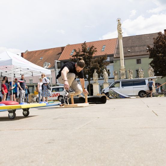 Ein Mann nimmt an einem Kegelspiel im Freien teil, mit Zuschauern, einem Zelt und Statuen im Hintergrund.