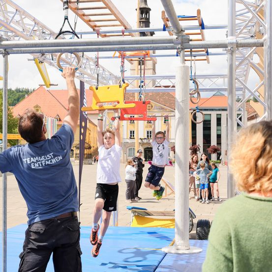 Eine Gruppe von Kindern und Erwachsenen nimmt an einem vergnüglichen Workout in einem Outdoor-Hindernisparcours teil. Ein Kind springt zwischen den Ringen.