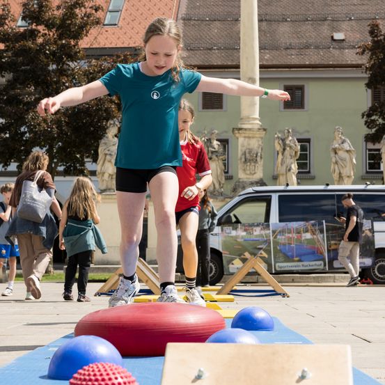 Ein Mädchen springt über einen roten Ball, umgeben von anderen im Spielplatz. Hinter ihr gehen Menschen an einem Van vorbei, mit einer Skulptur im Hintergrund.