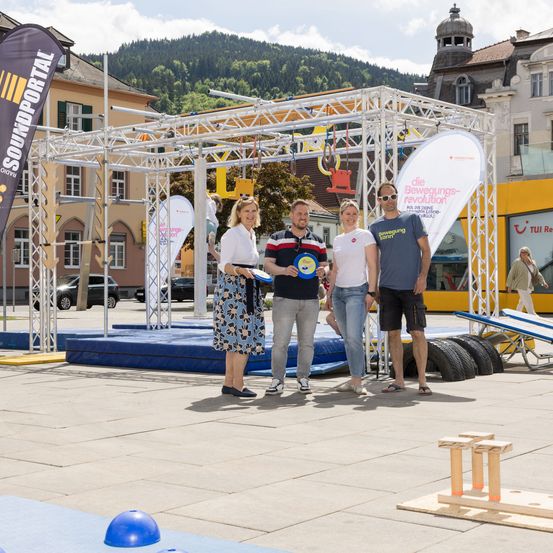 Eine Gruppe von Menschen steht auf einer Bühne mit Hindernissen in einem Außenbereich. Sie lächeln und posieren für ein Foto. Dahinter befindet sich eine Metallstruktur mit einem Banner und einem Gebäude. Vor dem Gebäude sind auch Autos geparkt.