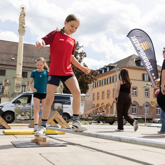 Ein Mädchen balanciert auf einem schmalen Balken auf einem Stadtplatz, mit einem Van und Gebäuden im Hintergrund. Andere Kinder und Erwachsene sind ebenfalls anwesend, einer von ihnen geht.