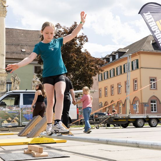 Ein junges Mädchen balanciert auf einem Balken draußen, trägt ein grünes T-Shirt und schwarze Shorts. In der Nähe sind Menschen und Gebäude zu sehen.