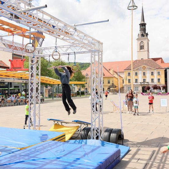 Eine Person führt eine Turnübung auf einem Outdoor-Hindernisparcours auf einem Stadtplatz vor, während Zuschauer zuschauen und eine Kirche im Hintergrund zu sehen ist.