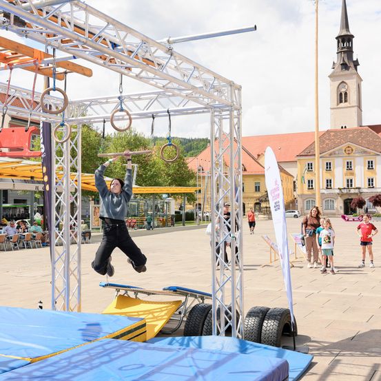 Eine Frau führt einen Klimmzug an einem Balken auf einem Stadtplatz durch, während Zuschauer zusehen. Im Hintergrund befinden sich ein Turm und Gebäude.