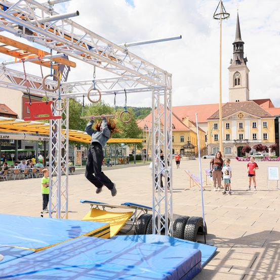 Eine Frau führt einen Stunt an einer Metallkonstruktion in einer Außenplaza vor. Im Hintergrund schauen Menschen und Kinder zu. In der Ferne befindet sich ein Gebäude mit einem Turm und einer Turmuhr.