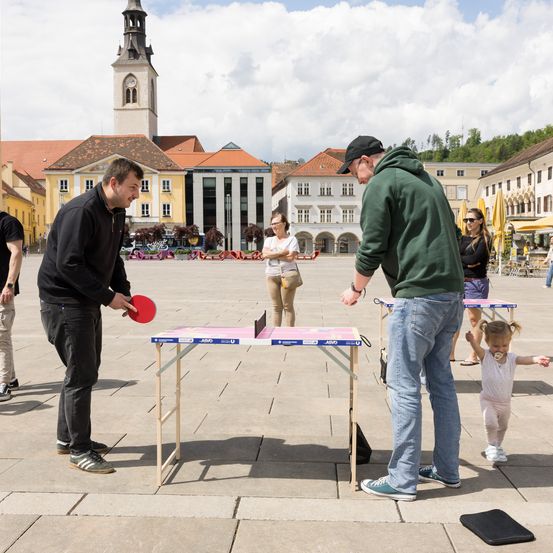 Zwei Männer spielen Tischtennis auf einem tragbaren Tisch auf einem Stadtplatz. Andere Leute sind in der Nähe, einschließlich einer Frau und eines Kindes. Im Hintergrund befinden sich Gebäude.