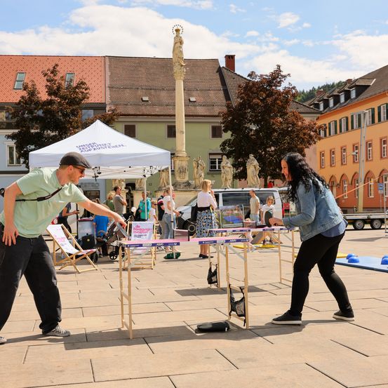 Ein Mann und eine Frau spielen draußen Tischtennis, mit einem Zelt und einer Statue im Hintergrund.