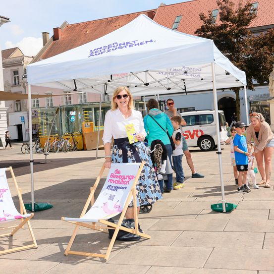 Eine Frau steht unter einem Vordach mit der Aufschrift 'Unser Zukunft Ausstellen' und einem Schild über 'Bewegungs-revolution'. In der Nähe versammeln sich Menschen, einschließlich Kinder. Ein Van und Fahrräder sind im Hintergrund zu sehen.