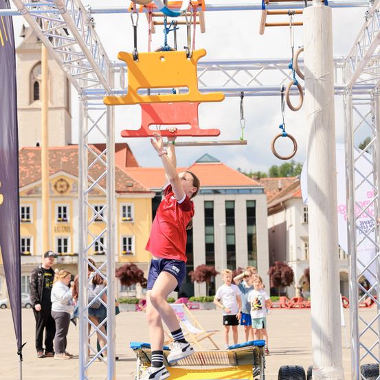 Eine junge Frau führt einen Stunt auf einem Hindernisparcours durch und greift ein gelbes Gerät, während Zuschauer und Gebäude im Hintergrund stehen.