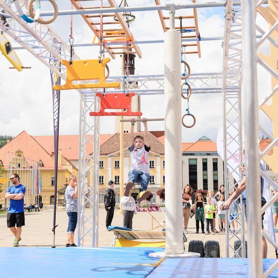 Ein junger Junge schwingt an einem roten Schaukelsitz, der an einer Metallkonstruktion in einem öffentlichen Park befestigt ist. Zuschauer beobachten von unten, einige stehen auf blauen Matten. Gebäude mit roten Dächern sind im Hintergrund.