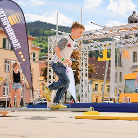 Ein Junge springt auf einer gelben Plattform in einem Outdoor-Hindernisparcours, mit einem Banner und einem Gebäude im Hintergrund.