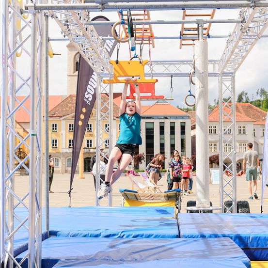 Ein junges Mädchen führt einen Stunt auf einem aufgehängten Hindernisparcours auf einem Stadtplatz durch. Gebäude und Menschen sind im Hintergrund.