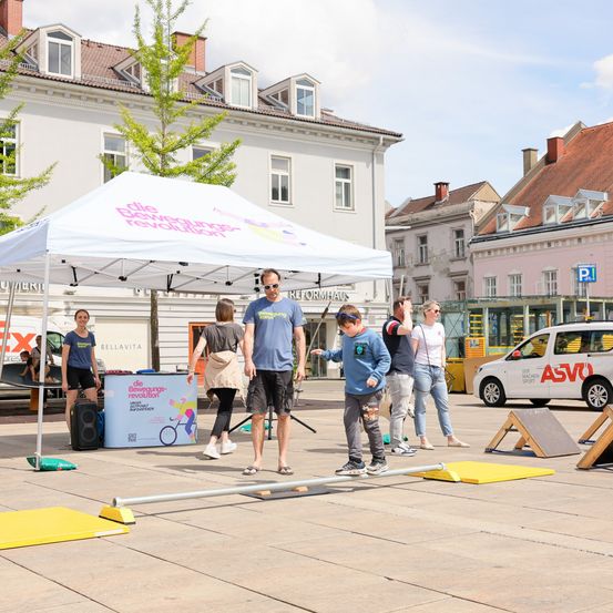 Eine Gruppe von Menschen, darunter ein junger Junge, nehmen an einer Veranstaltung unter einem weißen Zelt teil. In der Nähe ist ein weißer Van am Straßenrand geparkt.