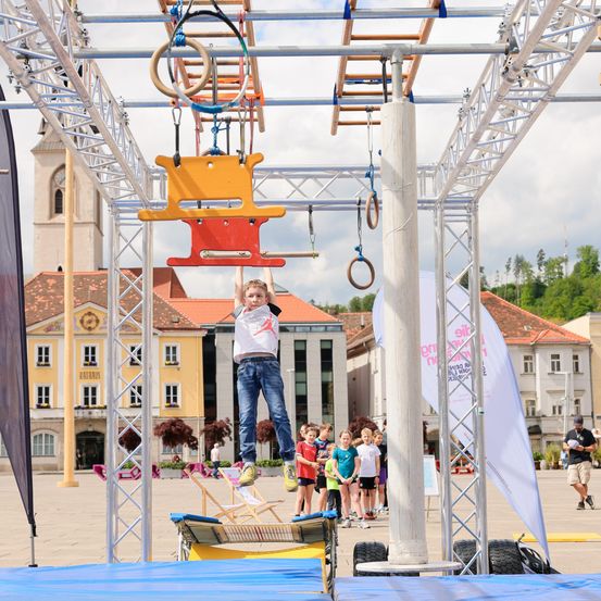 Ein junges Kind klettert auf einem bunten Hindernisparcours auf dem Stadtplatz, mit Gebäuden und einem Turm im Hintergrund.