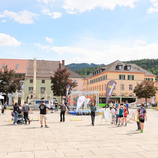 Menschen schlendern durch einen Stadtplatz mit mehreren Gebäuden und Statuen, einige spielen auf einem Hindernisparcours. Der Himmel ist blau mit Wolken.