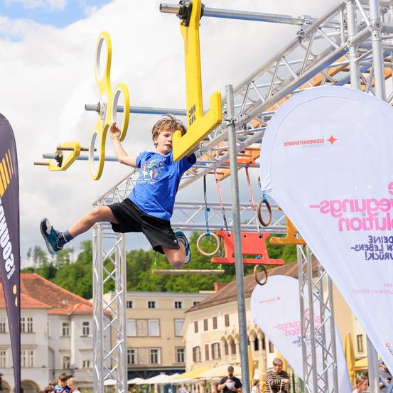 Ein junger Junge springt und hält sich an einem gelben Objekt in einem Hindernisparcours fest. Dahinter befinden sich mehrere Gebäude mit einigen Bäumen.