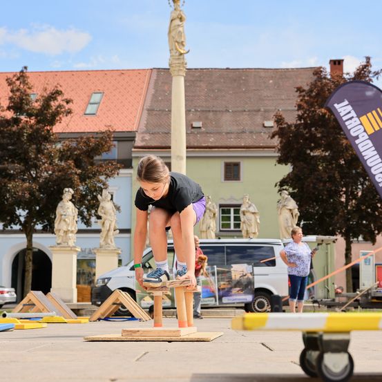 Eine junge Frau balanciert auf Holzklötzen auf einem Stadtplatz, mit Statuen und einem Gebäude im Hintergrund.