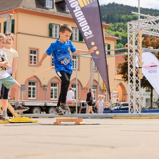 Ein junger Junge in einem blauen Fußballtrikot springt über eine Hürde im Park. Ein paar Kinder beobachten. Es gibt Gebäude, Bäume und einen Lastwagen im Hintergrund.