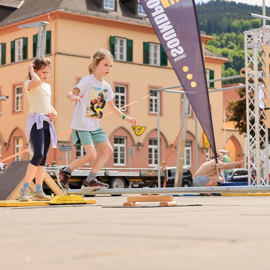 Zwei Mädchen absolvieren einen Hindernisparcours im Freien. Ein Mädchen sieht nach unten, während das andere läuft. Ein Banner und einige Holzblöcke liegen auf dem Boden.