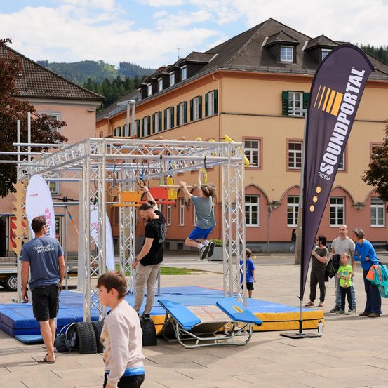 Eine Gruppe von Menschen spielt auf einem Hindernisparcours im Freien. Ein Junge klettert auf ein Netz, während andere zusehen. Ein Mann steht neben einer Fahne.