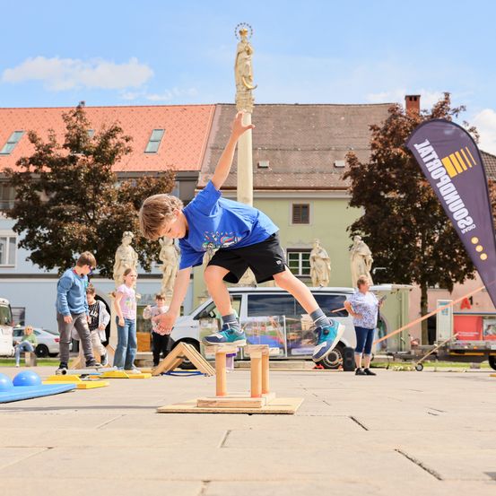 Ein Junge springt auf eine kleine Holzstruktur, während andere Kinder zusehen. Sie sind draußen in der Nähe einer Statue, Gebäuden und einem Banner.