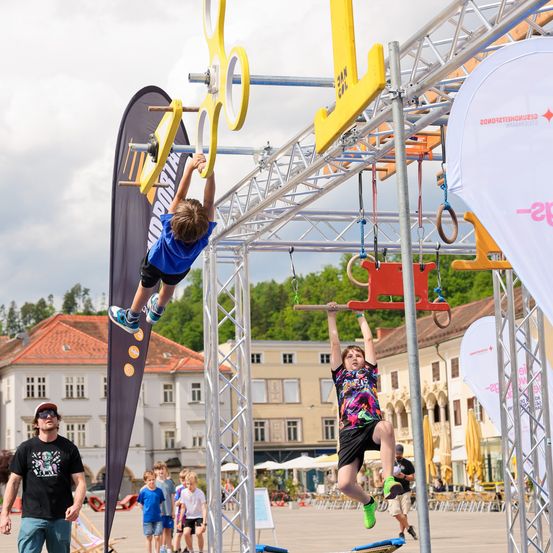 Zwei Kinder machen einen Hindernisparcours auf einer Freiluft-Plaza. Ein Junge hängt an einer gelben Stange, während ein anderer an einer roten Stange schwingt. Gebäude und Bäume sind im Hintergrund.