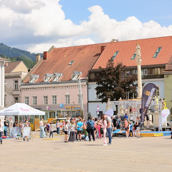 Ein Stadtplatz mit einer Menschenmenge, einem weißen Zelt und einem Gebäude mit rotem Dach. In der Mitte steht ein Denkmal mit einer Statue.