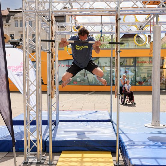 Ein Mann führt Klimmzüge an einer Stange bei einer Outdoor-Veranstaltung durch. Er trägt ein graues T-Shirt und schwarze Shorts. In der Nähe beobachtet eine Frau im Rollstuhl, und ein Gebäude ist im Hintergrund.