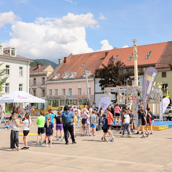 Viele Menschen versammeln sich auf einem Stadtplatz, einige in Sportkleidung, mit einem Zelt, einer Statue und Gebäuden im Hintergrund.