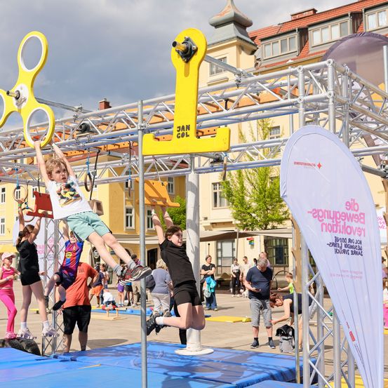 Eine Gruppe von Kindern und Erwachsenen beteiligt sich an einem Spielplatz-Hindernisparcours mit gelben Strukturen und Bannern. Ein Kind klettert auf einem gelben Objekt, während andere zuschauen. Im Hintergrund befinden sich Gebäude und Bäume.