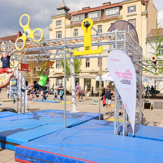 Kinder spielen auf einer Kletterstruktur in einem öffentlichen Außenbereich, umgeben von Gebäuden und Menschen. Ein Banner ist an der Metallstruktur montiert.