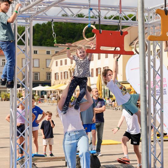 Mehrere Kinder spielen auf einer Metallkonstruktion mit Ringen, ein Mädchen wird von einem Erwachsenen hochgehoben. Dahinter stehen einige Leute und beobachten. In der Ferne ein Gebäude mit vielen Fenstern.